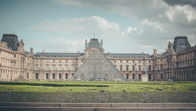 The image portrays the Louvre Museum with its iconic glass pyramid entrance in the center, framed by the classical architecture of the surrounding buildings. The sky is cloudy, creating a serene and majestic atmosphere. A well-manicured garden with hedges appears in the foreground.