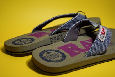 A pair of flip-flops with a patterned fabric strap and a textured sole is placed on a solid yellow background. The footbed has various graphics and logos printed on it in shades of dark blue and pink.