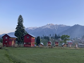 Two red wooden houses with blue roofs stand on a grassy field with a backdrop of snow-capped mountains. A few trees, including one tall evergreen, surround the area. Small wooden cabins are visible on the right side, and a clear sky hovers above.