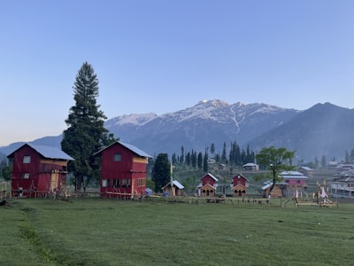 Two red wooden houses with blue roofs stand on a grassy field with a backdrop of snow-capped mountains. A few trees, including one tall evergreen, surround the area. Small wooden cabins are visible on the right side, and a clear sky hovers above.