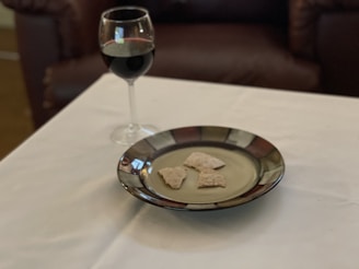 A glass of red wine and a plate with pieces of bread are placed on a white tablecloth. The background features a dark brown leather sofa, adding a cozy and relaxed atmosphere to the scene.