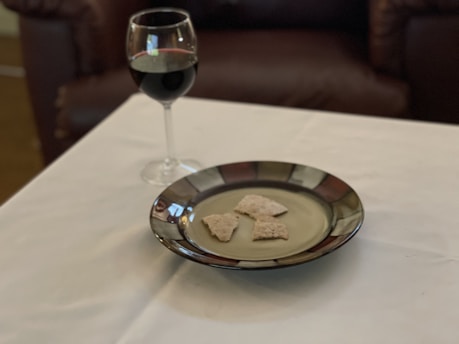 A glass of red wine and a plate with pieces of bread are placed on a white tablecloth. The background features a dark brown leather sofa, adding a cozy and relaxed atmosphere to the scene.