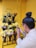 A person with glasses and a tattoo is taking a photo of an arrangement of flowers using a smartphone. The flowers are in a vertical stand against a bright yellow wall, with various colors including yellow, orange, and pink blossoms.