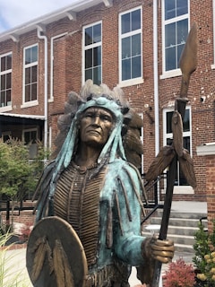A bronze statue depicting a Native American warrior standing in front of a brick building. The warrior is dressed in traditional attire with a headdress and holds a staff and shield adorned with feather motifs. The building features large rectangular windows and a gray staircase leading to an entrance.