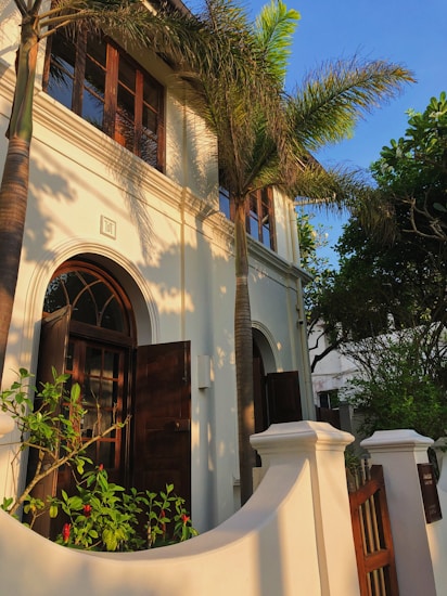 An elegant building with white walls and large arched windows framed by dark wooden shutters. Tall palm trees surround the structure, casting shadows on the facade. There is a small garden in front with lush green foliage and red flowers. A wooden gate and curved fence complete the entrance, creating a serene and tropical atmosphere.