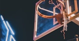 A basketball hoop and backboard illuminated by lighting, with a partially frayed net. The backdrop is a dimly lit environment, highlighting the bright hoop and the neon-like streaks of light next to it.