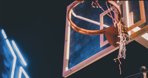 A basketball hoop and backboard illuminated by lighting, with a partially frayed net. The backdrop is a dimly lit environment, highlighting the bright hoop and the neon-like streaks of light next to it.
