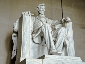 A large marble statue of a seated man, with a contemplative expression. The figure is dressed in a formal suit, sitting on a grand chair with one hand draped over the armrest. The craftsmanship highlights detailed features, conveying a sense of authority and reflection.