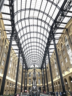 An architectural space with a high arched glass ceiling supported by black metal beams. The surrounding structure is made of brown brick with multiple levels of windows. In the center, there is a large, intricate metal sculpture. People are walking and standing around the area.