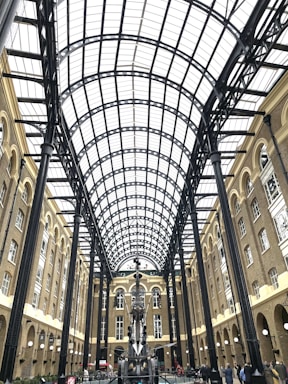 An architectural space with a high arched glass ceiling supported by black metal beams. The surrounding structure is made of brown brick with multiple levels of windows. In the center, there is a large, intricate metal sculpture. People are walking and standing around the area.