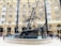 A large, intricate metal sculpture displays a combination of gears, wheels, and abstract shapes, situated in the center of a plaza. People are walking and gathering around the structure, and it is surrounded by a classic brick building with arched windows and black metal columns.
