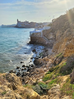 A scenic coastal view with rugged cliffs in the foreground, leading down to a rocky shoreline. The clear blue sea stretches into the distance, with waves lapping gently against the rocks. A town with historic buildings, including a notable church tower, is nestled against the shoreline, providing a picturesque backdrop. The sky is bright with sunlight creating a warm, inviting atmosphere.