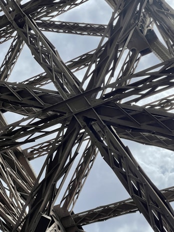 Intricate metallic structures form an elaborate network of beams and girders, creating an architectural design with geometric patterns. The beams appear sturdy and are set against a backdrop of a cloudy sky.