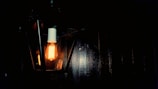 A vintage-style light bulb emits a warm glow, encased in a geometric lantern secured to a dark wall. The soft illumination contrasts with the surrounding darkness, accentuating the bulb's elongated filaments.