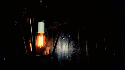 A vintage-style light bulb emits a warm glow, encased in a geometric lantern secured to a dark wall. The soft illumination contrasts with the surrounding darkness, accentuating the bulb's elongated filaments.