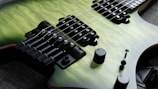 A close-up view of an electric guitar with a quilted green body finish. The image highlights the strings, pickups, and bridge, showcasing details like tuning pegs and knobs. The guitar is resting on a dark fabric surface.