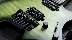 A close-up view of an electric guitar with a quilted green body finish. The image highlights the strings, pickups, and bridge, showcasing details like tuning pegs and knobs. The guitar is resting on a dark fabric surface.