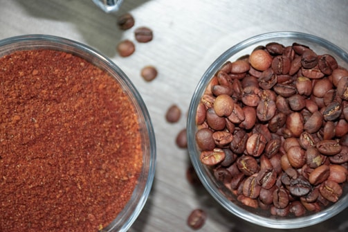 Two transparent bowls are placed on a surface. The left bowl contains finely ground coffee while the right bowl is filled with whole coffee beans. Several individual coffee beans are scattered across the surface between the bowls.