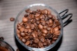 A clear glass cup filled with dark roasted coffee beans, placed on a wooden surface. The coffee beans have a shiny, oily texture, indicating freshness and quality.
