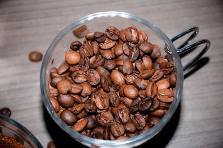 A clear glass cup filled with dark roasted coffee beans, placed on a wooden surface. The coffee beans have a shiny, oily texture, indicating freshness and quality.