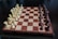 A wooden chessboard set up for a game with light-colored pieces on the left and dark-colored pieces on the right. The board has a checkered pattern, and the pieces are arranged in their starting positions, including pawns, rooks, knights, bishops, a queen, and a king for each color.