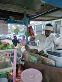 A street vendor is preparing food in a small roadside stall, surrounded by cooking utensils, pots, and packaged goods. In the background, a woman and a man sit on the sidewalk in front of parked cars and buildings.