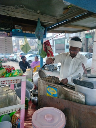 A street vendor is preparing food in a small roadside stall, surrounded by cooking utensils, pots, and packaged goods. In the background, a woman and a man sit on the sidewalk in front of parked cars and buildings.