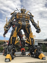 A large robot sculpture assembled from various metallic parts, prominently featuring yellow and black colors. It stands outdoors against a partly cloudy sky, with details resembling mechanical and automotive components.