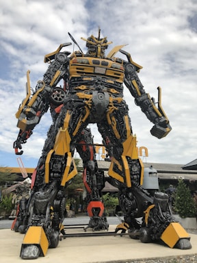 A large robot sculpture assembled from various metallic parts, prominently featuring yellow and black colors. It stands outdoors against a partly cloudy sky, with details resembling mechanical and automotive components.