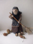 A handmade doll dressed in rustic clothing is holding a simple broom made of sticks and fibers. The doll's face is concealed with a cloth, resembling traditional attire. Acorns are scattered on the surface around the doll.