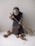 A handmade doll dressed in rustic clothing is holding a simple broom made of sticks and fibers. The doll's face is concealed with a cloth, resembling traditional attire. Acorns are scattered on the surface around the doll.