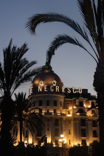A grand, historic hotel adorned with a prominent, illuminated sign reading 'Le Negresco' sits elegantly surrounded by tall palm trees. The building's façade is bathed in warm, golden lighting that highlights its classical architecture, including a large dome on top. The sky is a dusky blue, suggesting early evening.