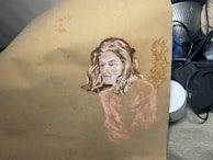 A painting on a piece of paper depicting the portrait of a person with light-colored hair, blending into the background. The artwork features soft brush strokes and a muted color palette, with additional items like a metal strainer and some compact containers partially visible in the background.