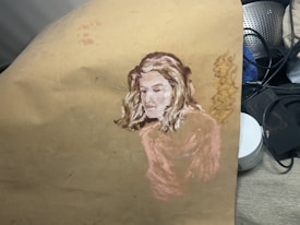 A painting on a piece of paper depicting the portrait of a person with light-colored hair, blending into the background. The artwork features soft brush strokes and a muted color palette, with additional items like a metal strainer and some compact containers partially visible in the background.