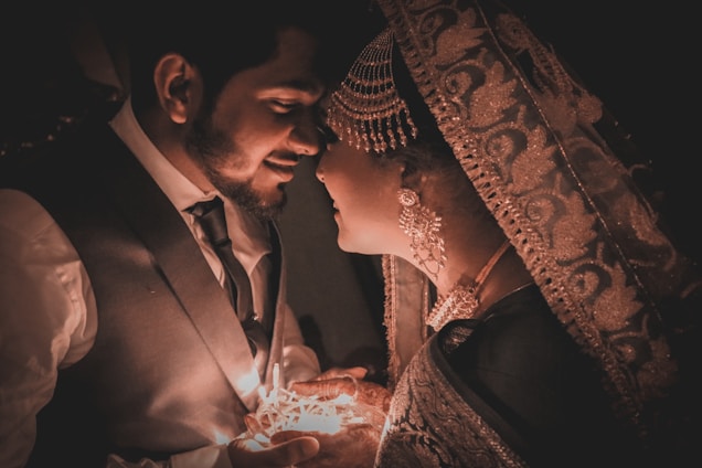 A couple is facing each other closely, illuminated by a soft, warm light. The man is wearing a suit and tie, while the woman is adorned with traditional ornate jewelry and a richly embroidered veil. They hold a small source of light between them, creating a romantic atmosphere.