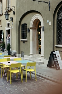 A quaint street scene with colorful outdoor tables and chairs in pastel shades, placed along a cobblestone pathway. The facade of a building includes an arched doorway adorned with lights and a window with iron bars. A person stands in the doorway, talking on a phone. A chalkboard sign outside advertises offerings of wine, snacks, and coffee.