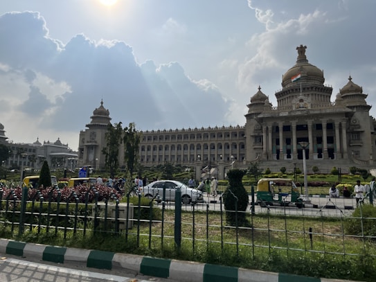 The image depicts a grand, historic building with a domed structure and intricate architectural details. Flags are visible on top of the building, indicating its importance. In front of the building, there are well-maintained gardens, a fenced area, and several people and vehicles including cars and auto-rickshaws. The sky above is partly cloudy with sun rays piercing through.