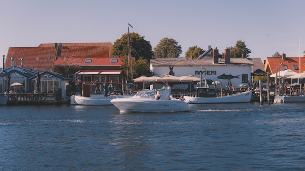 A scenic waterfront view featuring several small buildings with red-tiled roofs and outdoor seating areas. Boats are docked at the wooden piers, and a white speedboat is cruising across the water. Trees in the background and a clear blue sky suggest a pleasant day.