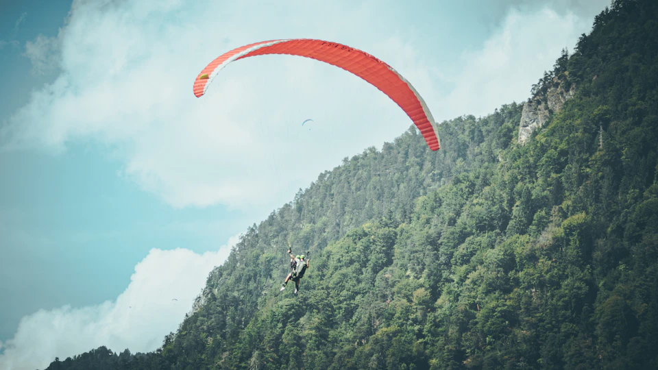 The image depicts a person paragliding over a lush green mountain landscape, with their body suspended in mid-air and attached to a vibrant red parachute that is billowing against the backdrop of the clear blue sky dotted with fluffy white clouds. The paraglider appears to be soaring high above the verdant trees and rocky terrain below.

The perspective of the image is from below, looking up at the airborne adventurer, creating a sense of awe and admiration for their daring feat.