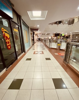 A shopping mall corridor with tiled floors and shops lining both sides. The corridor has a geometric pattern on the floor and a skylight in the ceiling. Shops on the left include a reflexology store, while others are further down the corridor. Metal railings and a well-lit atmosphere complete the scene.