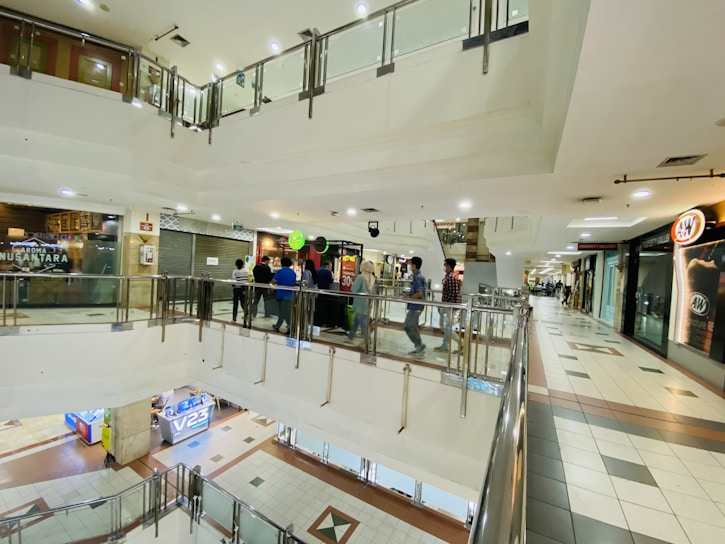 A multi-level indoor shopping mall with people walking along a corridor on an upper floor. There are several shops with closed shutters and a fast-food restaurant sign visible. The floor is tiled, and the area is well illuminated with ceiling lights.