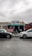 A small fashion shop with a corrugated metal roof and a sign above the entrance displays various clothing items. The building, painted in various colors, is situated in an urban setting with a wet street and two parked cars in front. A person dressed in casual clothing walks past the shop.