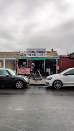A small fashion shop with a corrugated metal roof and a sign above the entrance displays various clothing items. The building, painted in various colors, is situated in an urban setting with a wet street and two parked cars in front. A person dressed in casual clothing walks past the shop.