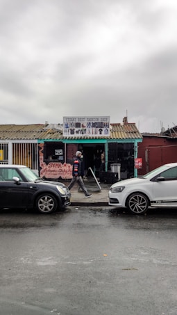 A small fashion shop with a corrugated metal roof and a sign above the entrance displays various clothing items. The building, painted in various colors, is situated in an urban setting with a wet street and two parked cars in front. A person dressed in casual clothing walks past the shop.