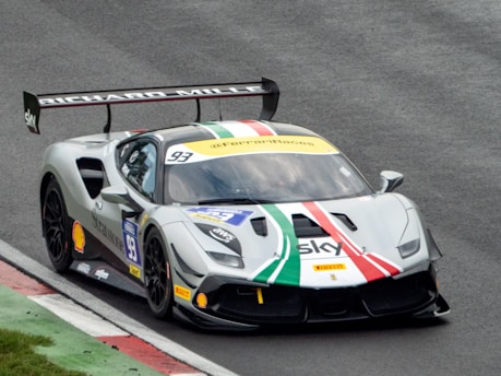 A racing car on a racetrack featuring a prominent rear wing and sponsor logos. The car has a design with white, green, and red stripes across its hood and roof. The track has a mixture of asphalt and painted curbs.