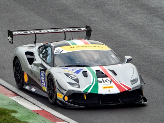 A racing car on a racetrack featuring a prominent rear wing and sponsor logos. The car has a design with white, green, and red stripes across its hood and roof. The track has a mixture of asphalt and painted curbs.