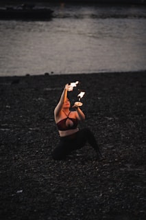 A person kneels on a dark, pebbled beach while performing a fire dance at dusk. The performance involves manipulating flaming objects with graceful movements. The surrounding scenery includes a body of water with a dimly visible shoreline and a boat in the background.
