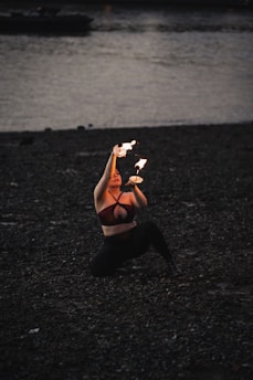 A person kneels on a dark, pebbled beach while performing a fire dance at dusk. The performance involves manipulating flaming objects with graceful movements. The surrounding scenery includes a body of water with a dimly visible shoreline and a boat in the background.