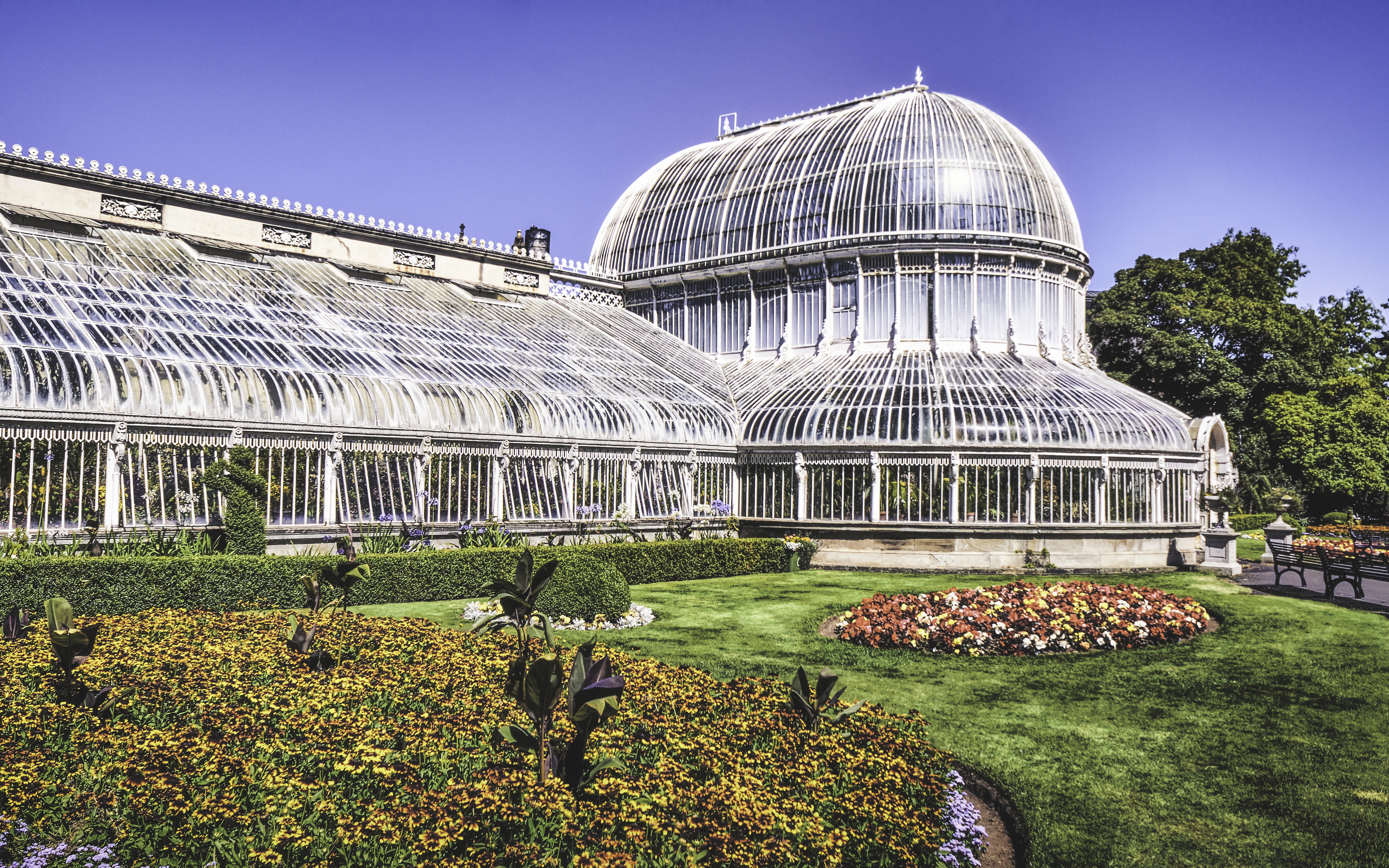 The Victorian glass Palm House at Botanic Gardens Belfast under blue sky with colourful flower beds.