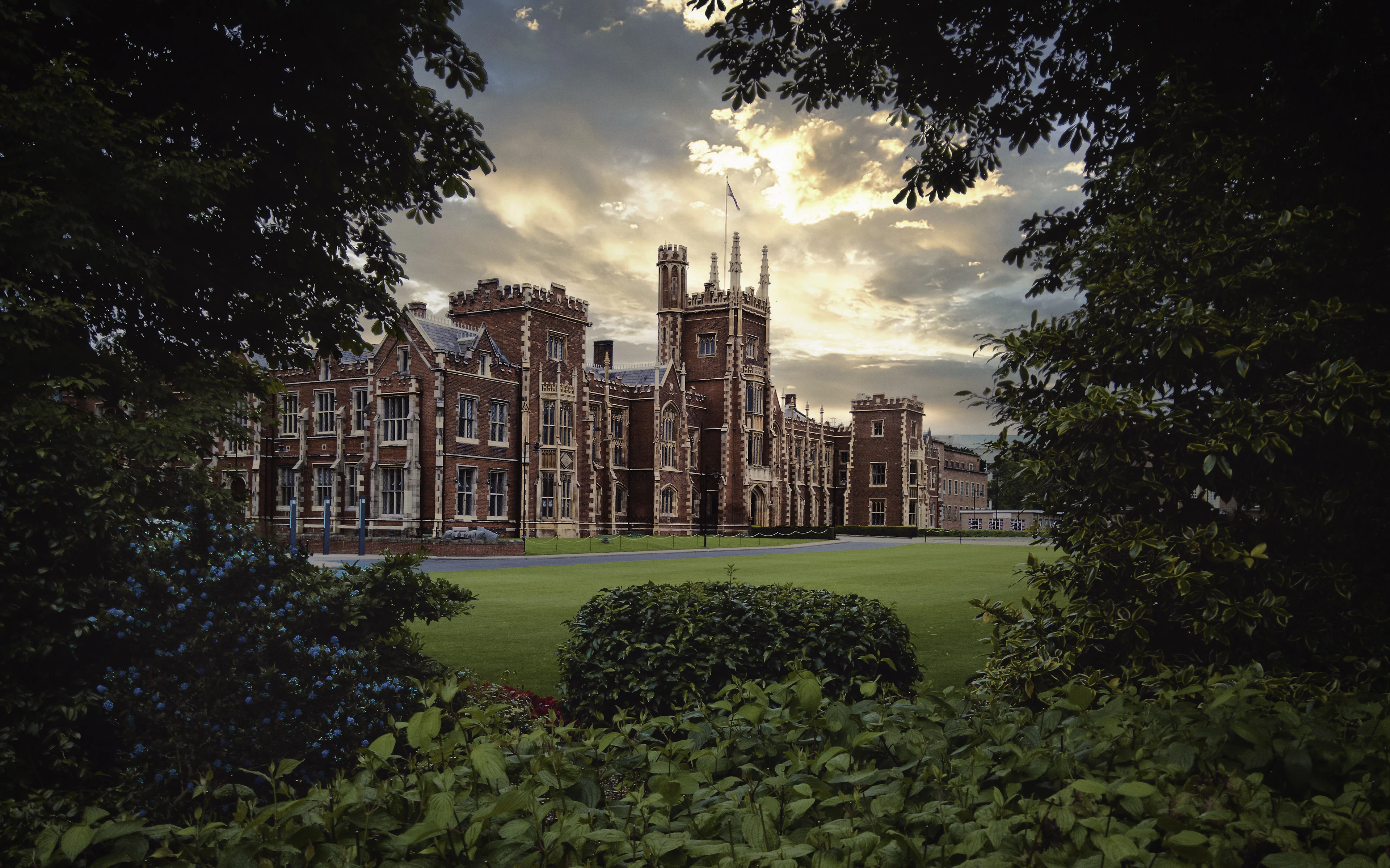 The red-brick Gothic Revival Lanyon Building at Queen's University Belfast across its lawn, framed by dark summer foliage, with evening sunlight breaking through gold-edged cloud behind its tower.